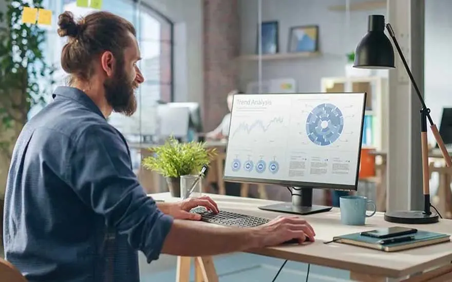 Een man met knot werkt aan een modern bureau, bestudeert een monitor vol grafieken en diagrammen met het label trendanalyse.
