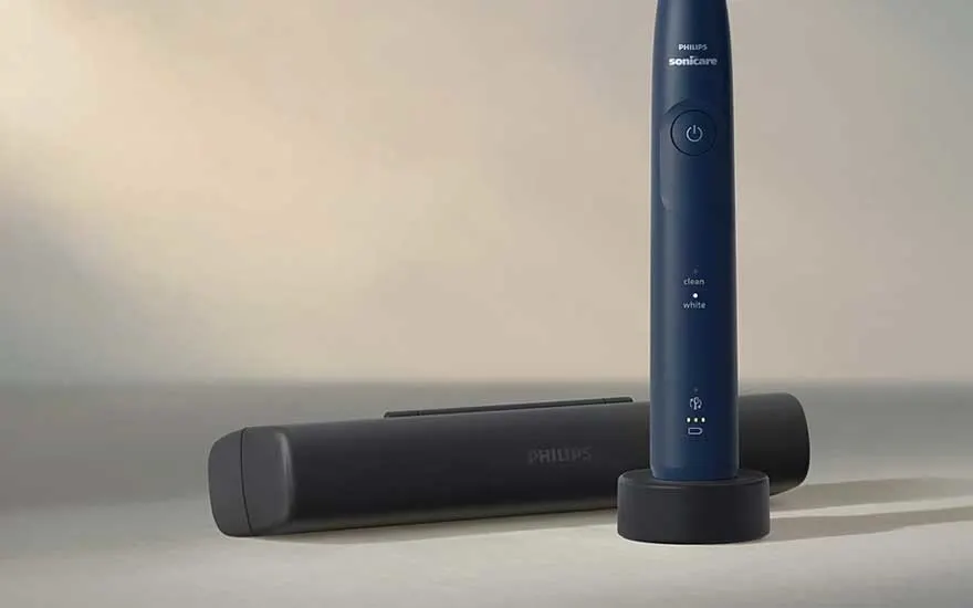 A blue Philips Sonicare stands upright on its charger, with a closed travel case beside it in soft, warm light.