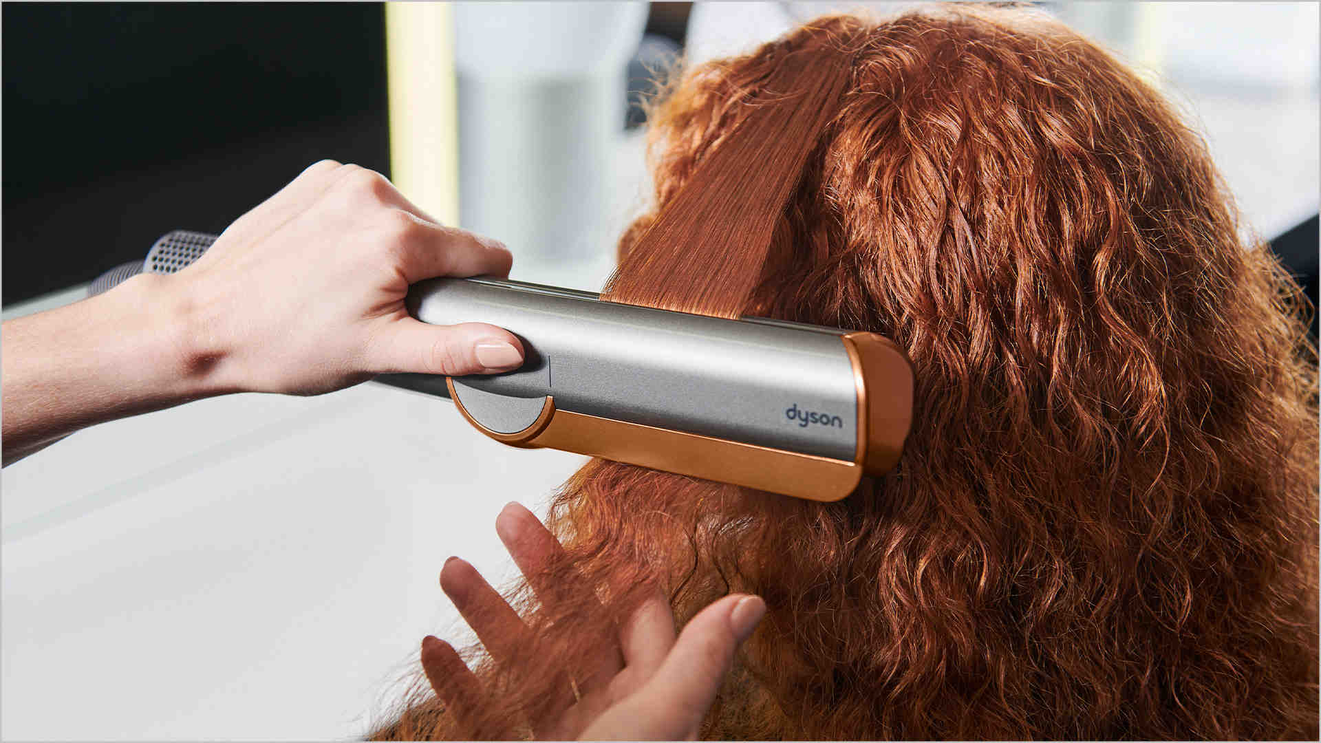 Een grijze Dyson stijltang wordt gebruikt op rood haar, met een hand die het haar vasthoudt. De achtergrond is vaag.