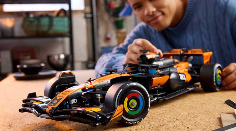 Man in blauwe trui bouwt aan een oranje LEGO Formule 1-auto op tafel, met bril, koffiekop en gezellige kamer eromheen.