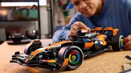 Man in blauwe trui bouwt aan een oranje LEGO Formule 1-auto op tafel, met bril, koffiekop en gezellige kamer eromheen.