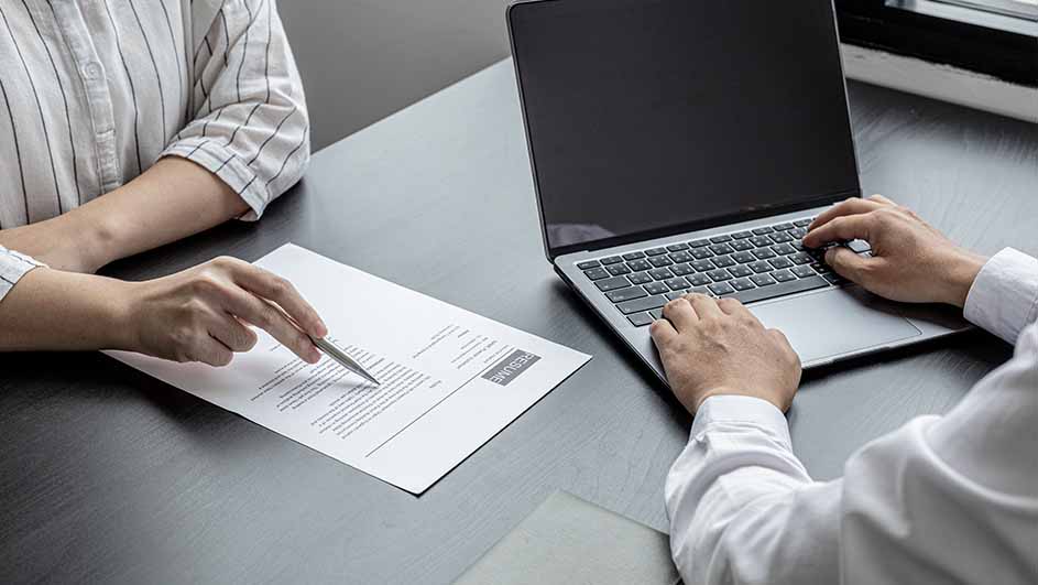 Op een grijze tafel zit iemand met een pen bij een cv en iemand anders typt op een laptop.