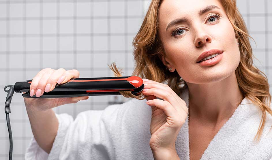 Een vrouw in een witte badjas stylet een haarlok met een rood-zwarte stijltang voor een betegelde achtergrond.