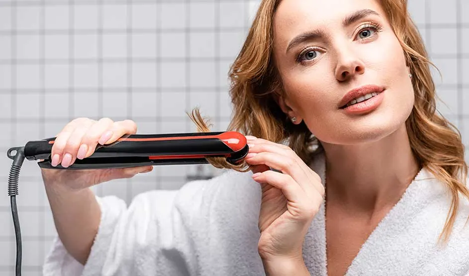 A woman in a white bathrobe styles a strand of hair with a red-and-black straightener against a tiled background.