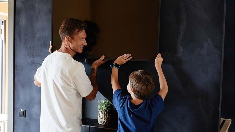A man and a boy are installing a television on a wall. A plant sits on a shelf below the television.