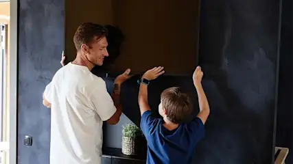 A man and a boy are installing a television on a wall. A plant sits on a shelf below the television.