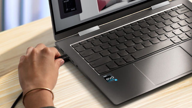 Een hand steekt een zwarte USB-stekker in een grijze laptop op een houten tafel. Het scherm is gedeeltelijk zichtbaar.