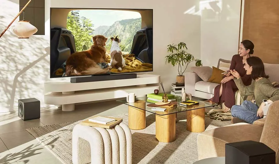 Two women relax while watching a large television showing dogs, in a bright and modern living room.