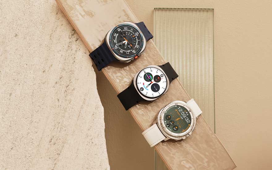 Drie smartwatches met verschillende bandjes liggen op een lichte plank, met sportieve en klassieke wijzerplaten zichtbaar.