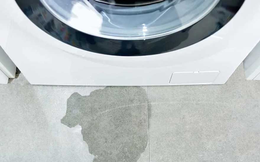 A white washing machine with a glass door and black rim. In front of the appliance, a large puddle of water is visible on the grey tiled floor, indicating a water leak or overflow from the machine.