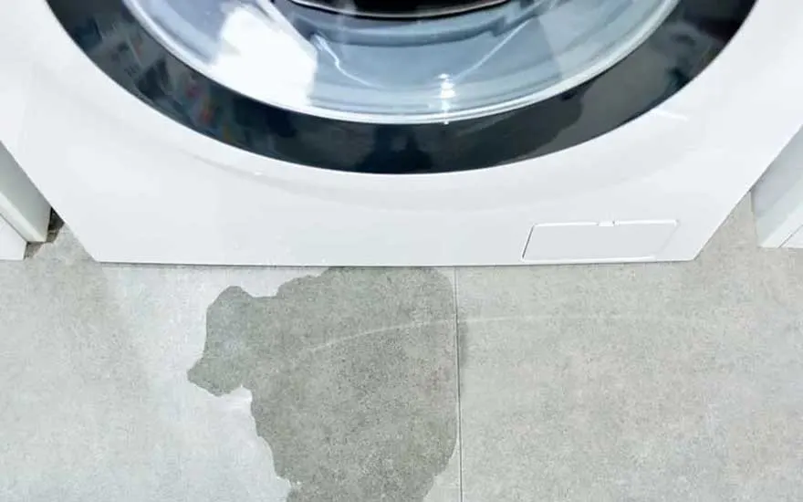 A white washing machine with a glass door and black rim. In front of the appliance, a large puddle of water is visible on the grey tiled floor, indicating a water leak or overflow from the machine.