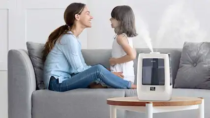 Een moeder en dochter zitten samen op een bank, glimlachen naar elkaar naast een luchtbevochtiger die stoom afgeeft op de salontafel.