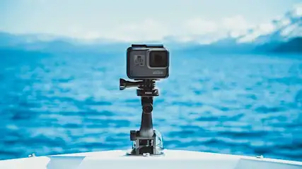 A gray action camera is mounted on a boat, facing the viewer, with a blue sea and mountains in the background.