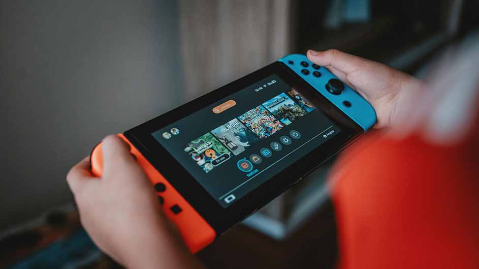 Handen houden een Nintendo Switch vast met blauwe en oranje controllers, het scherm toont spelpictogrammen.