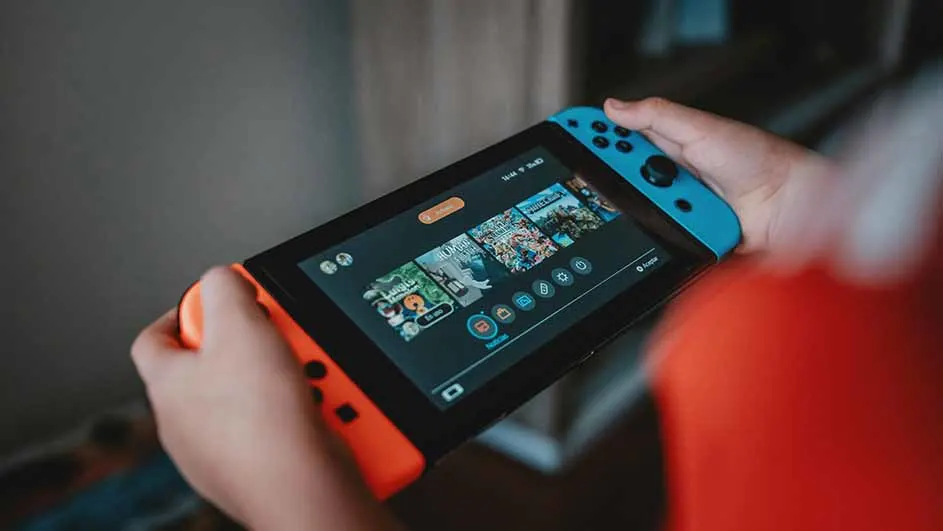 Handen houden een Nintendo Switch vast met blauwe en oranje controllers, het scherm toont spelpictogrammen.