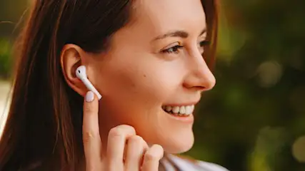 Een vrouw glimlacht met een oortelefoon in haar oor en een groene, wazige achtergrond.