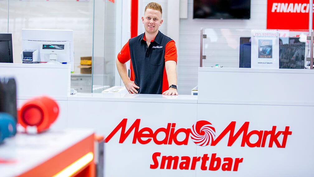 Een medewerker in rood-zwart uniform staat achter een witte MediaMarkt Smartbar balie. Kassa en schermen zijn zichtbaar.