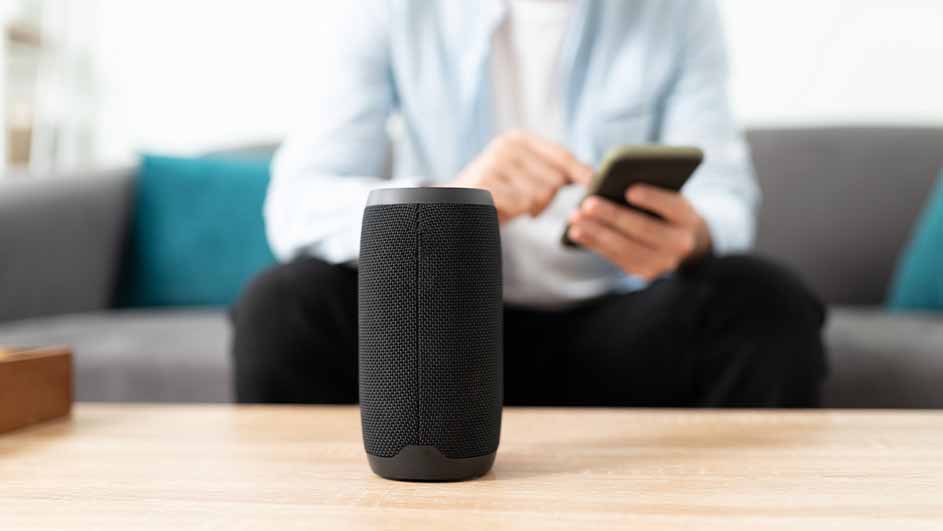 Een zwarte speaker staat op een tafel. Op de achtergrond zit een persoon op een grijze bank met kussens en gebruikt een telefoon.