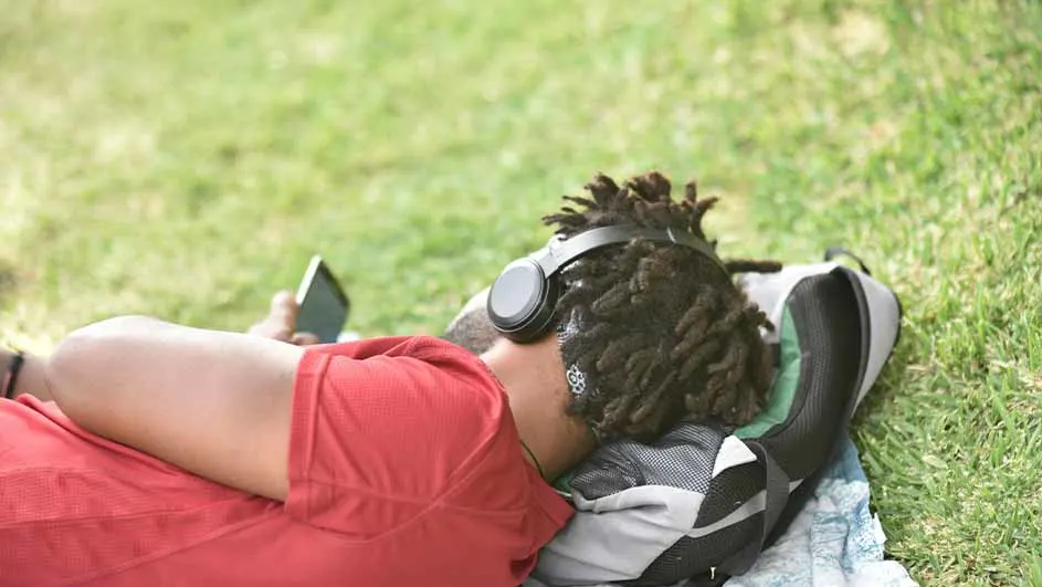 Een persoon met een rood shirt, koptelefoon en telefoon ligt op een tas in het gras.