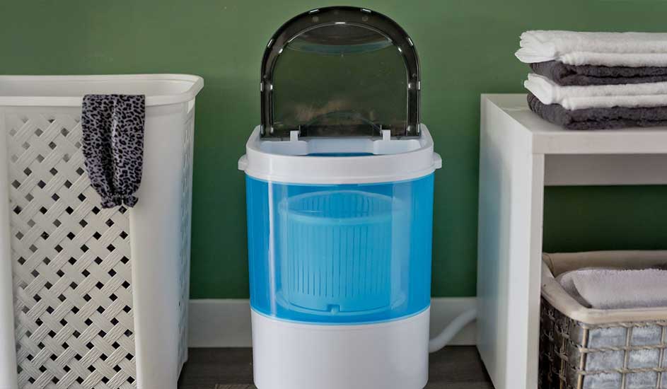 Een compacte, blauwe mini-wasmachine zonder zichtbaar merk staat tussen een wasmand en een wit rek met handdoeken in een wasruimte.