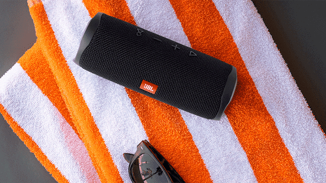 Een zwarte JBL-luidspreker ligt op een gestreepte oranje-witte handdoek. Onder de handdoek ligt een zwarte zonnebril.