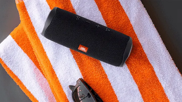 Een zwarte JBL-luidspreker ligt op een gestreepte oranje-witte handdoek. Onder de handdoek ligt een zwarte zonnebril.