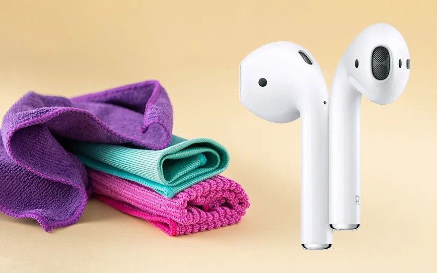 Two white wireless earbuds stand upright beside neatly folded microfibre cloths in bright purple, turquoise and pink on a beige background.