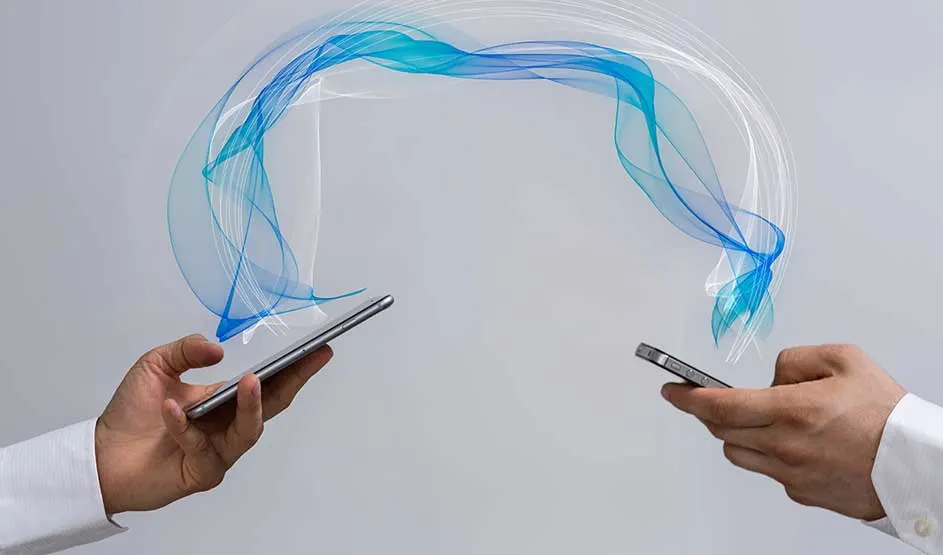 Twee handen houden smartphones; een blauwe golf verbindt beide toestellen en symboliseert draadloze gegevensoverdracht. Geen merk zichtbaar.