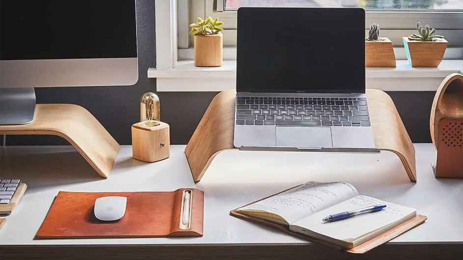 Een bureau met een laptop op een houten standaard, een notitieboek met een pen, en kantooraccessoires.