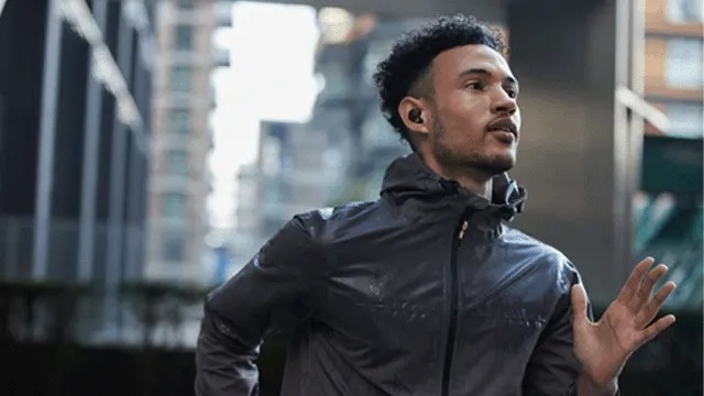 Man in a grey running jacket jogs through the city, wearing small black wireless earbuds and looking focused as he moves past tall buildings.