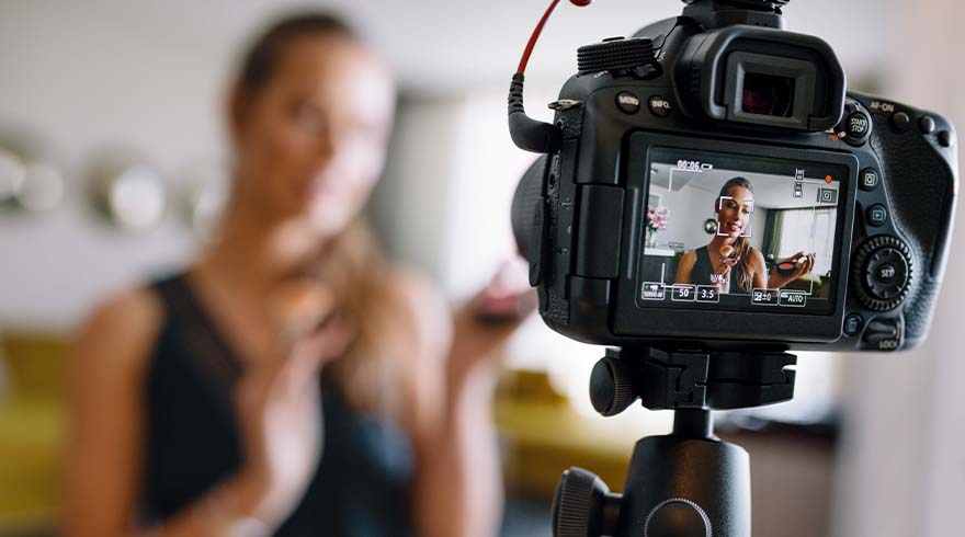 Een camera op een statief filmt een vrouw die in de verte staat. De camera toont een opname van dezelfde vrouw.