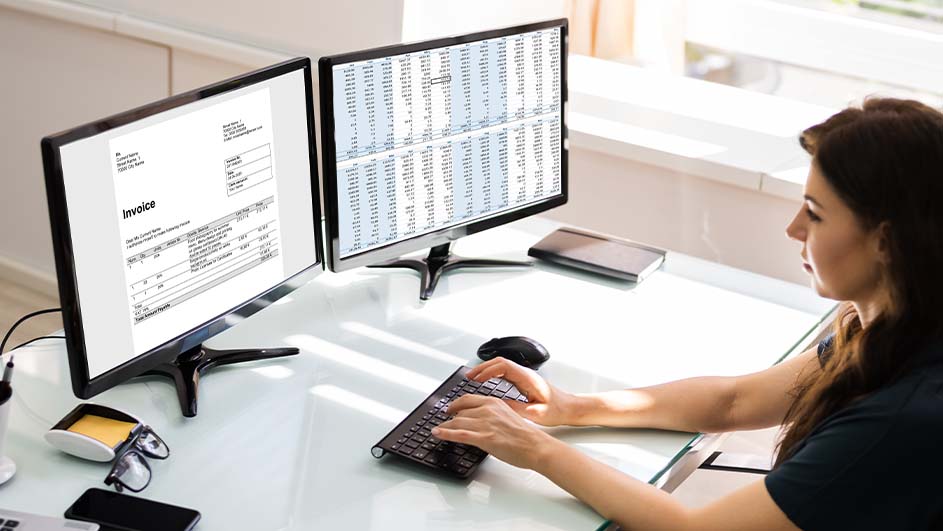 Vrouw typt op toetsenbord voor twee monitoren; één toont een factuur, de ander een spreadsheet. Bril en telefoon op bureau.
