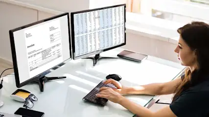Vrouw typt op toetsenbord voor twee monitoren; één toont een factuur, de ander een spreadsheet. Bril en telefoon op bureau.