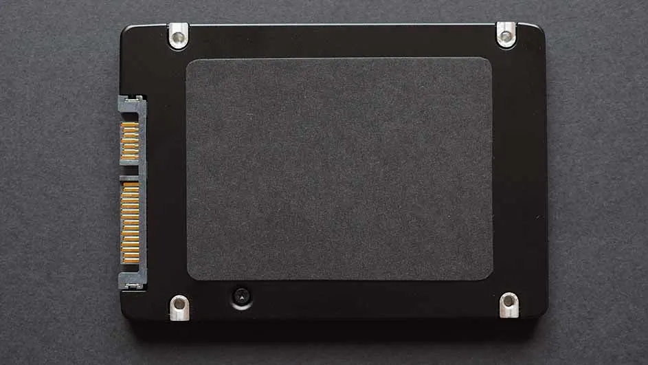 Een zwarte SSD met een donkergrijs label ligt op een grijze achtergrond, met gouden pinnen aan de linkerzijde en vier zilveren bevestigingspunten.
