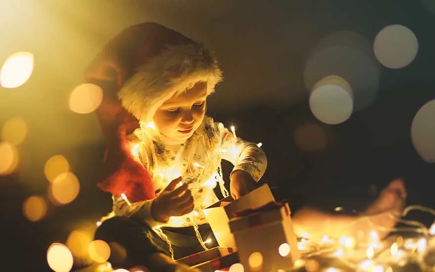 Een klein kind met een kerstmuts zit in het donker tussen fonkelende lichtjes en opent nieuwsgierig een cadeaudoos vol warme gloed.