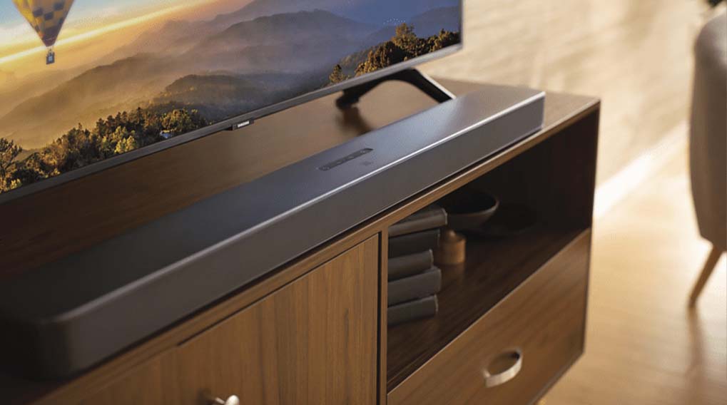 Een donkergrijze soundbar staat op een houten tv-meubel onder een tv met een berglandschap en een luchtballon.