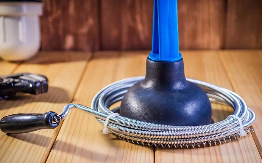 A black rubber plunger with blue handle rests on a coiled metal drain snake on a wooden floor. A tool is visible to the left. The background shows a brown wooden wall and a white container.