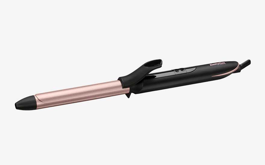 Slim rose gold curling iron with black handle and clamp rests diagonally on a light background, designed to create neat, shiny curls and waves.