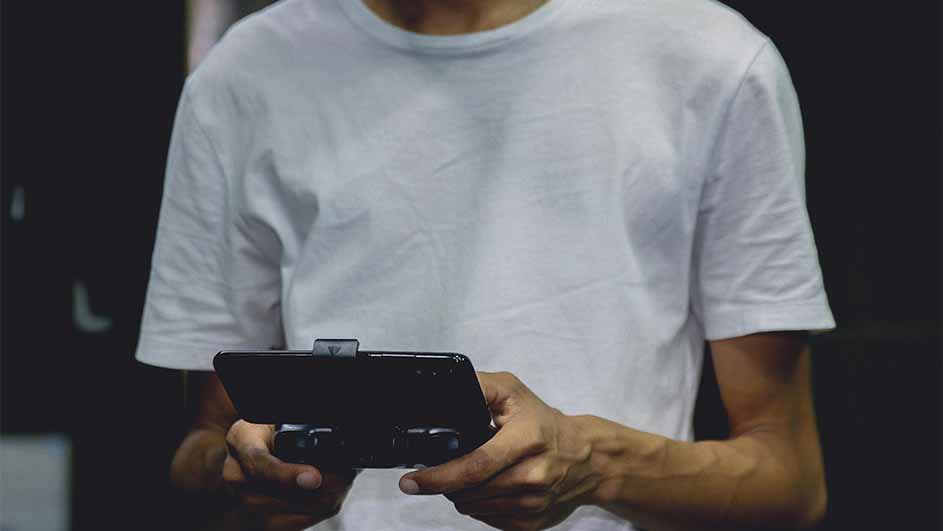 Een persoon met een wit T-shirt houdt een zwarte telefoon met een gamepad-accessoire vast tegen een donkere achtergrond.