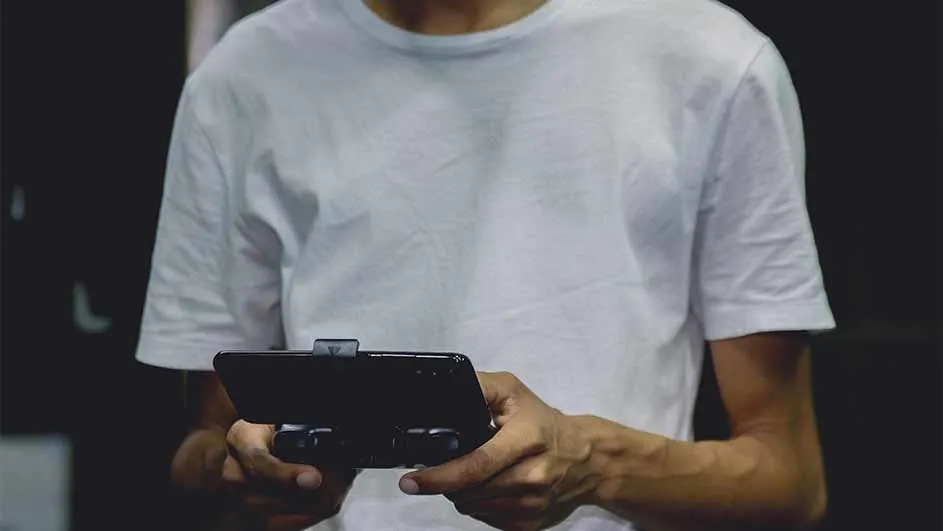Een persoon met een wit T-shirt houdt een zwarte telefoon met een gamepad-accessoire vast tegen een donkere achtergrond.