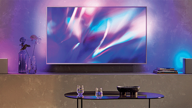 Een tv met abstracte afbeelding hangt boven een kast met decoraties, in een paars verlichte kamer.