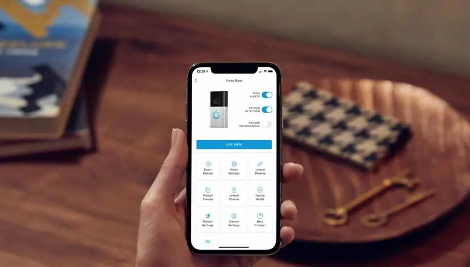 Hand houdt een smartphone met een app voor een slimme deurbel. Boeken, een dienblad en decoratieve items zijn zichtbaar op tafel.