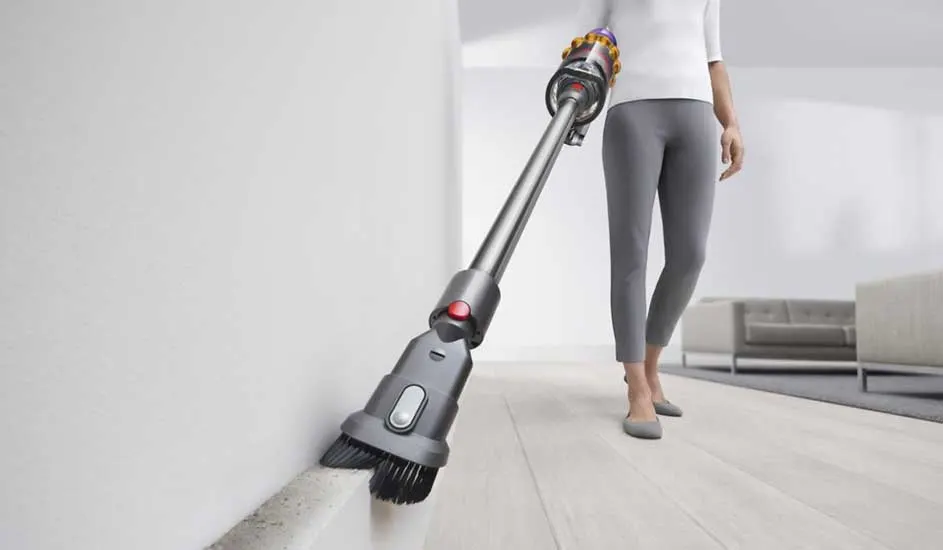 A woman vacuums the skirting board with a long cordless cleaner and soft brush, lifting fine dust neatly along the white wall.