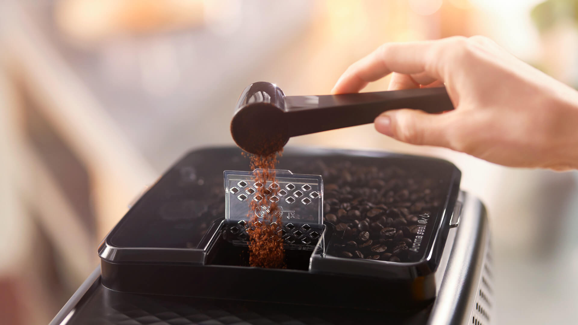 Koffiebonen worden met een zwarte lepel in een transparant bakje van een zwart apparaat gegoten.