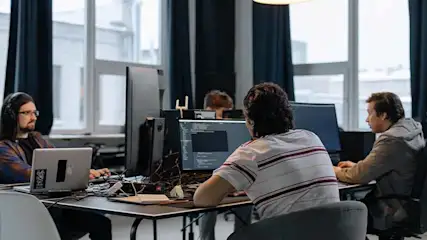 Kantooromgeving met werknemers achter computers aan bureaus, grote ramen en gordijnen op de achtergrond.