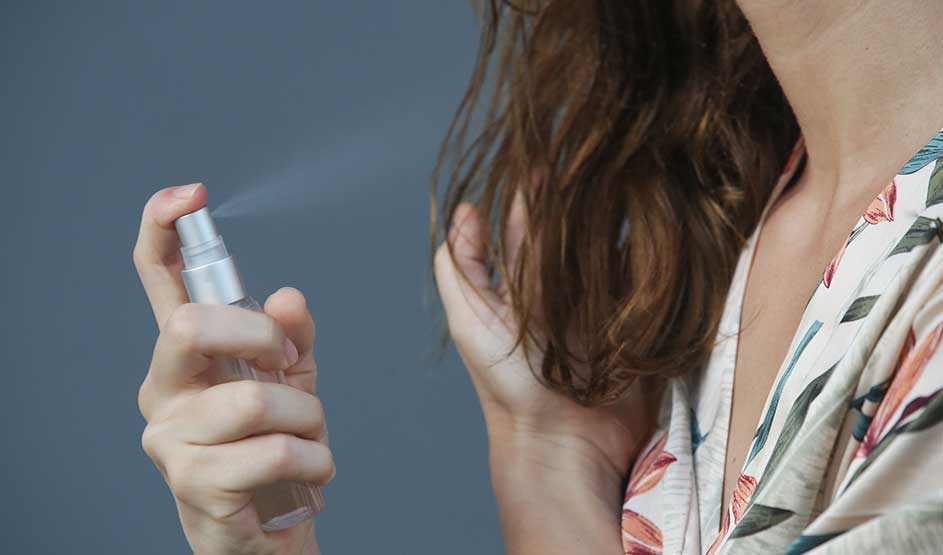 Iemand verstuift een verzorgende spray op nat haar, terwijl een hand de lokken bijeenhoudt tegen een rustige achtergrond.