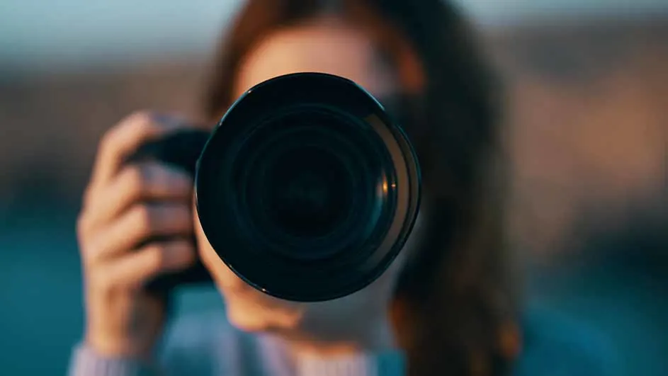 Een persoon houdt een camera met een grote lens vast, gericht naar voren. De achtergrond is onscherp met blauwe en oranje tinten.