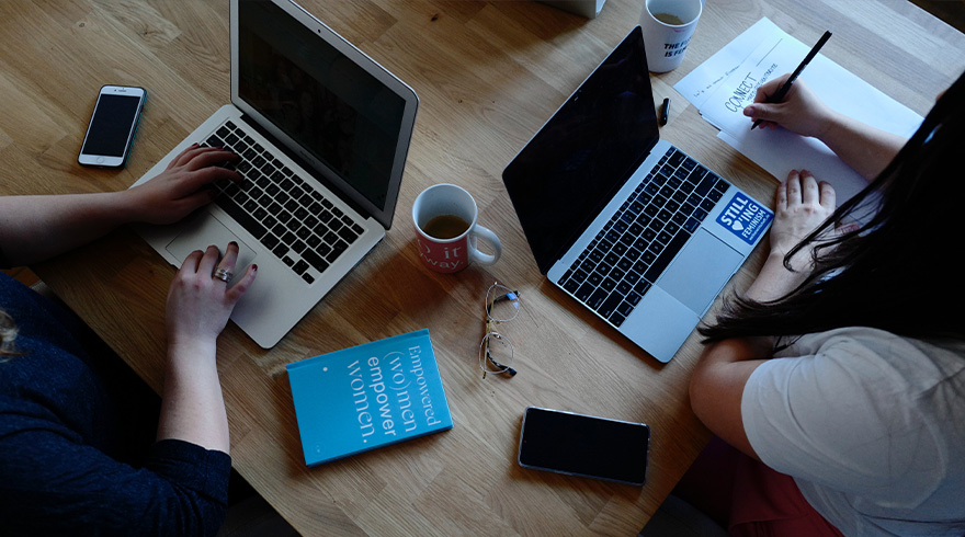 Twee personen werken aan laptops aan een houten tafel, met koffiekopjes, een boek, een telefoon en een bril.