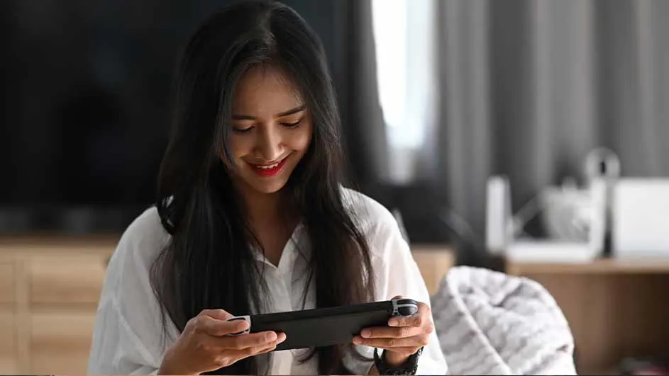 Een vrouw glimlacht en speelt op een zwarte handheld gameconsole in een kamer met gordijnen.