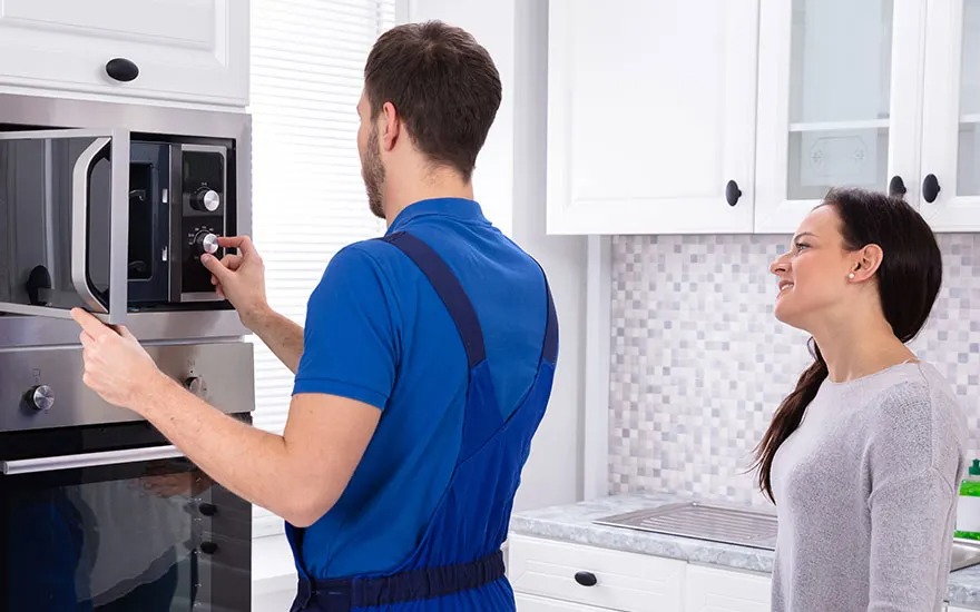 Monteur in blauwe overall stelt ingebouwde oven in moderne witte keuken af, terwijl een glimlachende vrouw toekijkt naast hem.